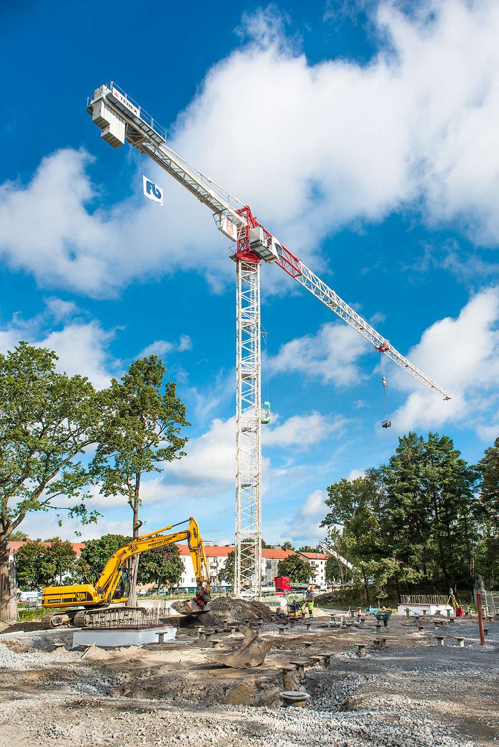 Tall tower crane with a construction site below, surrounded by trees and blue sky.